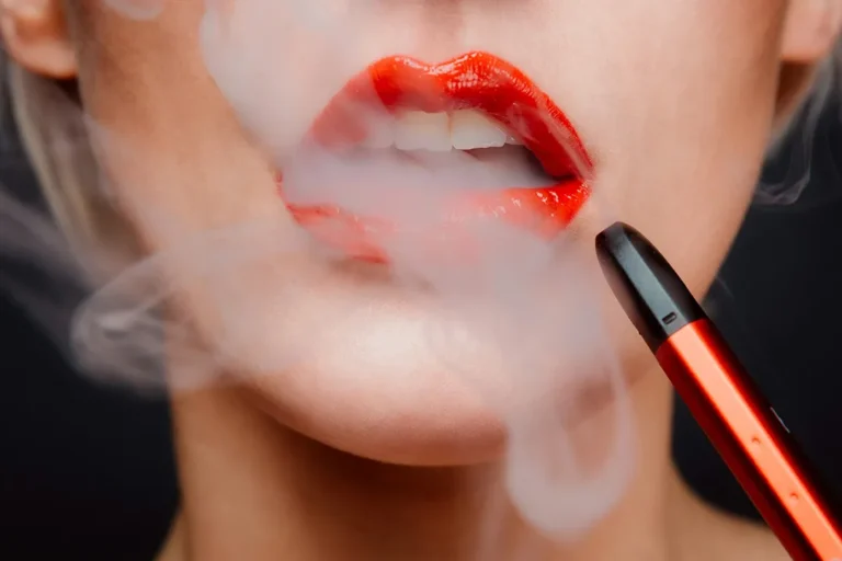 一幅女性特寫圖片,塗有鮮紅唇膏的嘴唇正呼出電子煙的白色霧化蒸汽,手中握著一支時尚的深橙紅色電子煙主機。此圖展示了現代替煙方案的使用場景。